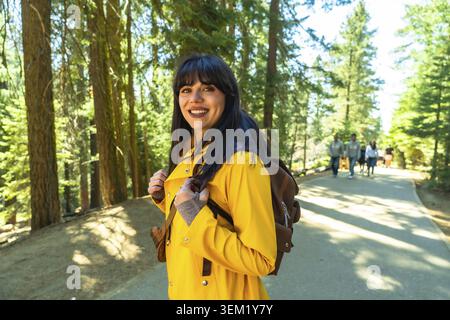 Young woman smiling and wearing a yellow raincoat and backpack, enjoying an outdoor adventure on an paved trail surrounded by tall trees in sequoia na Stock Photo