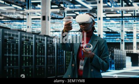 Data center system administrator uses VR headset gear and AR simulation to manage database storage rigs. Cloud computing server room worker uses virtual reality technology VFX visualization, camera B Stock Photo