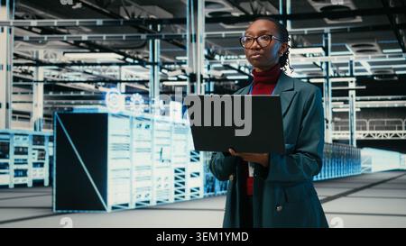 Female programmer walking in data center uses AR simulation and laptop to monitor systems. African american woman in server hub uses notebook to do optimizations, VFX compositing, camera A Stock Photo