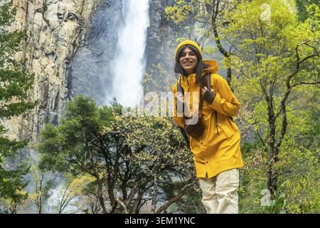 Smiling woman wearing a yellow raincoat and hat, carrying a backpack, standing on a trail near bridalveil fall in yosemite national park, surrounded b Stock Photo