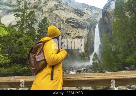 Woman wearing yellow rain jacket and beanie, carrying a backpack, standing on a wooden bridge enjoying the view of lower yosemite falls and rocky land Stock Photo