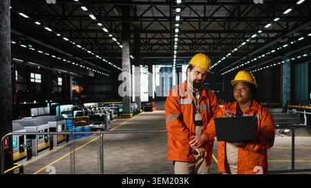 Diverse engineers, using walkie-talkie, portable transceiver, heavy industry plant, supervising welding stations and steel work with laptop in large-scale machinery hall. Manufacturing process. Stock Photo
