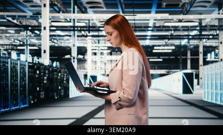 Woman in server farm using laptop to improve electronics optimizations. Female technician walking in data center using notebook to monitor system performance, identifying bottlenecks, camera A Stock Photo