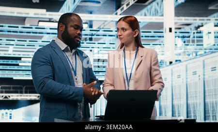 Data center coworkers collaborating to manage enterprise cloud computing IT solutions using laptop. Team of programmers discussing, monitoring futuristic server farm facility workspace, camera B Stock Photo