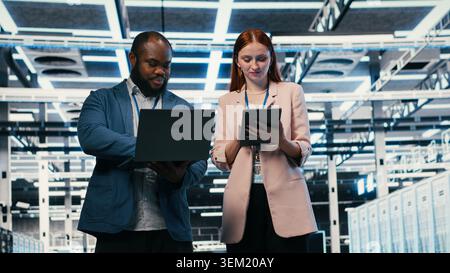 Optimizing electronics, laptop and team of software developers in server farm. Brainstorming, notebook and teamworking colleagues in data center monitoring system performance bottlenecks, camera B Stock Photo