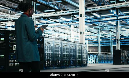 Female programmer walking in data center using tablet to write code, compiling software patches. African american woman in server hub using device application to run programming script, camera A Stock Photo