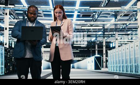 Optimizing electronics, laptop and team of software developers in server farm. Brainstorming, notebook and teamworking colleagues in data center monitoring system performance bottlenecks, camera A Stock Photo