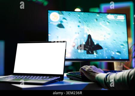 Isolated screen laptop next to man playing with friends online, shooting asteroids together in spaceship flying game. Mockup notebook next to gamer cooperating with teammates in coop videogame Stock Photo