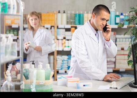 Male pharmacist talking by phone while working with computer in drugstore Stock Photo