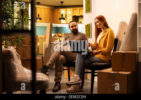 Homeowners using laptop for interior design inspiration and sitting among cardboard boxes and household items. Couple planning furniture layout and appliances during new house relocation. Stock Photo