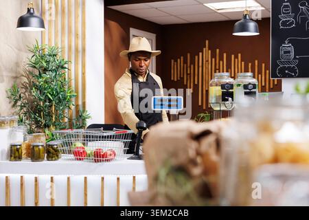 Focus on background of african american male shopkeeper at the checkout counter. Black man wearing a hat, arranging items over at the cashier desk in bio-store. Fresh, eco friendly produce displayed. Stock Photo