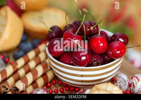 Macro view of a striped ceramic bowl overflowing with fresh red cherries on a holiday dessert table with blurred background pastries. Stock Photo