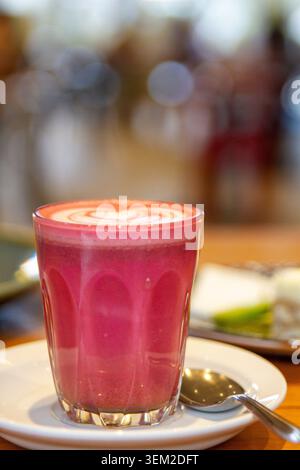 Close-up view of a pink beetroot latte hot drink featuring white foam heart latte art in a glass cup on a saucer with a silver spoon. Stock Photo