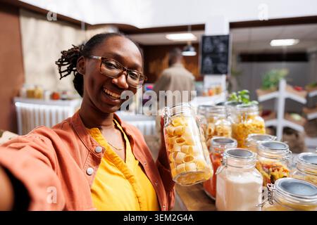 In bio-food shop, black woman wearing spectacles holds glass container loaded with spaghetti. Organic products, vlogging, digital camera, and customer videography are all examples of online marketing. Stock Photo