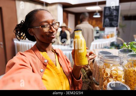 A young black woman films herself in a zero-waste shop promoting eco friendly grocery shopping. She highlights fresh, homegrown products and engages her online audience through vlogging. Stock Photo