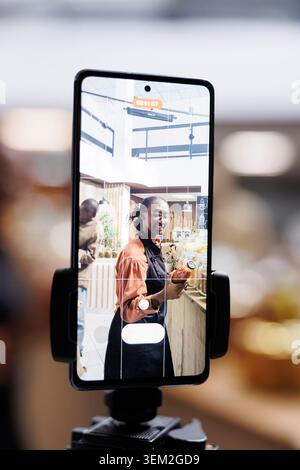 African American store owner uses cell phone and selfie stick for vlogging, promoting sustainable, healthy and natural items. Organic, fresh food in glass jars, appealing to eco-conscious customers. Stock Photo