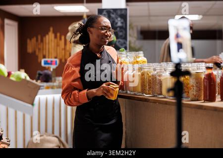 A black woman promotes eco friendly products in a modern supermarket. African american female owner filming a vlog using a smartphone and tripod stand, showcasing local, organic and sustainable items. Stock Photo