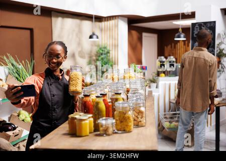African American female owner of a zero waste shop promotes eco friendly products. Filming a video call, she showcases fresh groceries, homegrown jars, and sustainable packaging. Stock Photo
