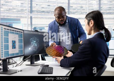 Multiracial engineering team inspecting silicon wafer substrates assisted by artificial intelligence at office desk. Teamworking colleagues use AI technology on PC to analyze semiconductor slice Stock Photo