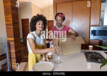 Diverse female friends unpacking kitchen island with cardboard box, vase, plates, coffee cups Stock Photo