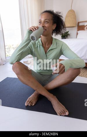 African American woman sipping from textured water bottle while cooling down on yoga mat in bedroom Stock Photo