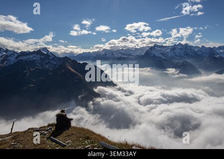 View of a person sitting on a grassy ridge at Stoos overlooking a thick sea of clouds and snow-covered mountain peaks under a bright blue sky in Morsc Stock Photo