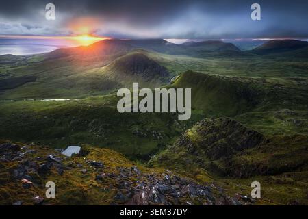 View of the Coumanare mountains under a dramatic sunset sky with clouds over green slopes and rocky terrain in County Kerry, Ireland. Stock Photo