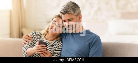Happy Caucasian Relaxing On Couch Together, Using Smartphone At Home, Lifestyle Concept Stock Photo