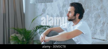 Thoughtful Young Man Sitting On Couch In Living Room, Pensive Mood, Healthcare Concept Stock Photo