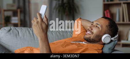 Smiling Young Black Man Enjoying Music On Smartphone While Lying On Couch At Home Stock Photo