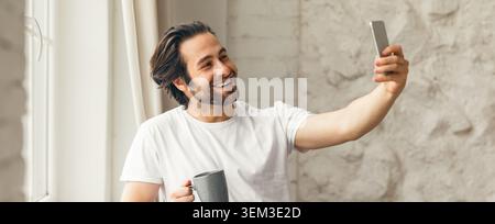 Happy Caucasian Man Taking Selfie While Enjoying Coffee In Modern Home Setting Stock Photo