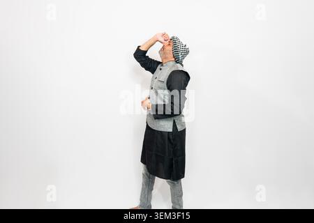 A man wearing traditional Indian clothing, including a black kurta and grey vest. He is posed with his hand raised to his forehead against a plain whi Stock Photo