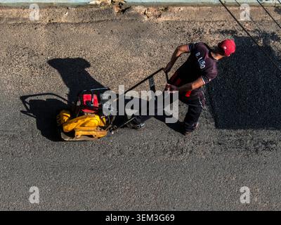 Marilia, SP, Brazil, June 03, 2026: A construction worker in uniform operates a yellow vibratory plate compactor machine during road maintenance and a Stock Photo