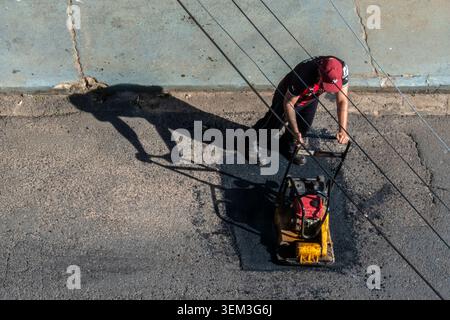 Marilia, SP, Brazil, June 03, 2026: A construction worker in uniform operates a yellow vibratory plate compactor machine during road maintenance and a Stock Photo