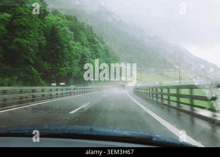 A point of view shot from inside a vehicle driving along an asphalt highway during a rainstorm, showing a wet road surface, traffic ahead, and lush fo Stock Photo