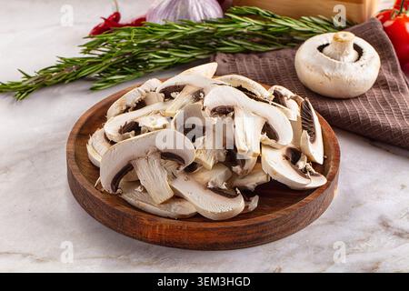 Fresh sliced champignon mushrooms arranged on round wooden cutting board with rosemary sprigs, garlic bulb, and red chili peppers on marble surface, n Stock Photo