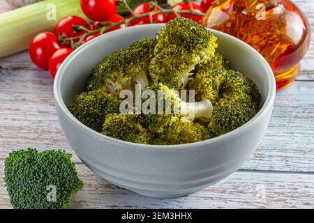 Steamed broccoli florets in a gray bowl with cherry tomatoes and oil on a wooden table Stock Photo