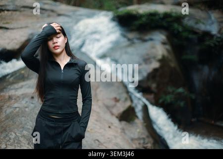 Young asian woman feeling exhausted and tired resting in nature near beautiful waterfall wearing black activewear enjoying outdoors after difficult hi Stock Photo