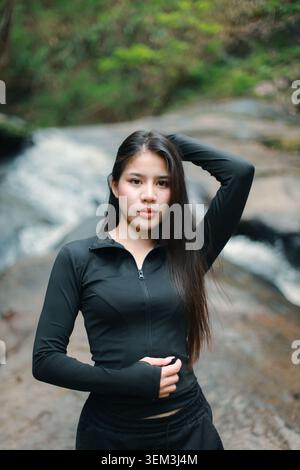 Confident young asian woman standing outdoor in peaceful nature wearing black activewear for forest portrait near river rocky stream looking deeply an Stock Photo