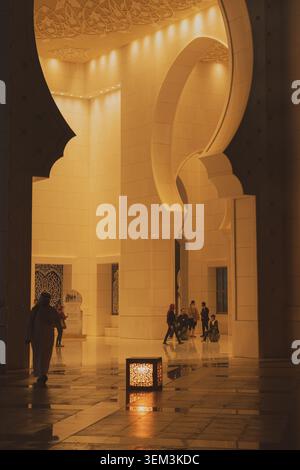 Warmly lit mosque interior with tall arches, polished floor, and small groups of visitors walking through a spacious hall Stock Photo
