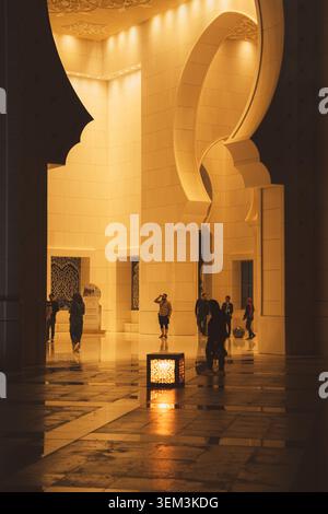 Warmly lit mosque interior with tall arches, reflective floor and people walking through the grand hall. Stock Photo