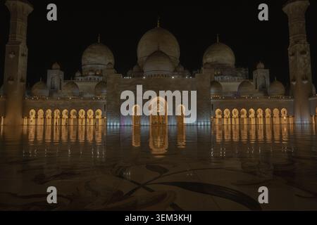 Illuminated mosque with domes and arches at night, reflected on a wet marble floor, creating a calm and symmetrical scene. Stock Photo