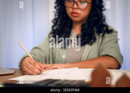 Young female student wearing glasses is focused on writing or drawing a project at her desk at night. Stock Photo