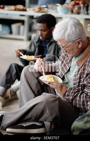 Senior Man Eating Meal at Humanitarian Aid Center with People in Need Stock Photo