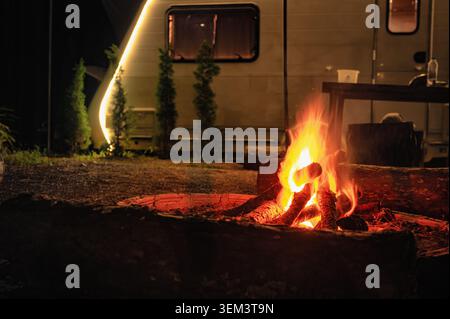 Glowing campfire surrounded by logs in front of a warmly lit camper, creating a cozy night camping vibe. Stock Photo