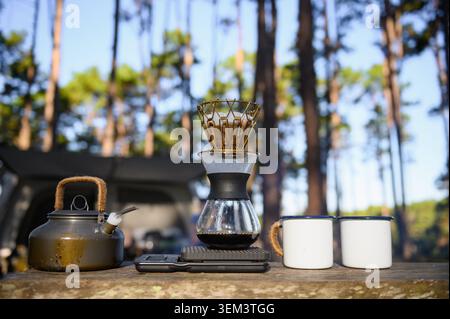Forest coffee setup with a kettle, a pour-over coffee maker, and two enamel mugs on a wooden table. Surrounded by trees and sunlight, it creates a pea Stock Photo