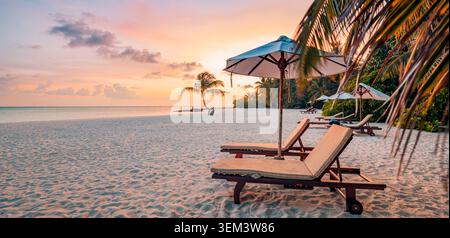 Romantic sunbeds umbrella on tropical beach at warm pastel sunset with palms. Panoramic golden sky ocean. Luxury honeymoon resort vacation travel art Stock Photo