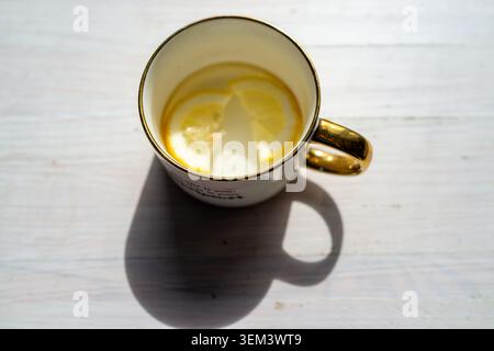 Top view of a ceramic cup holding a light lemon drink with citrus slices, placed on a pale wooden surface with strong shadow and bright natural daylight Stock Photo