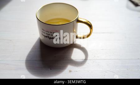 A white ceramic cup with a gold rim and handle sits on a bright table, filled with a warm yellow drink and casting a long soft shadow in natural sunlight Stock Photo