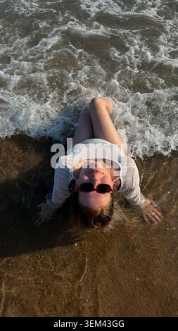 Young woman in sunglasses lying on sandy beach, partially submerged in ocean waves, enjoying sunny day with water splashing around her Stock Photo